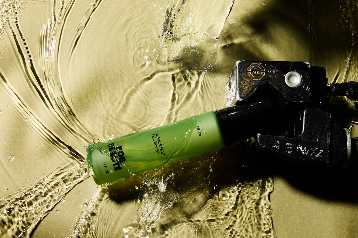 A translucent green BOB BEAUTÉ bottle with a black cap lying in rippling water on a golden surface