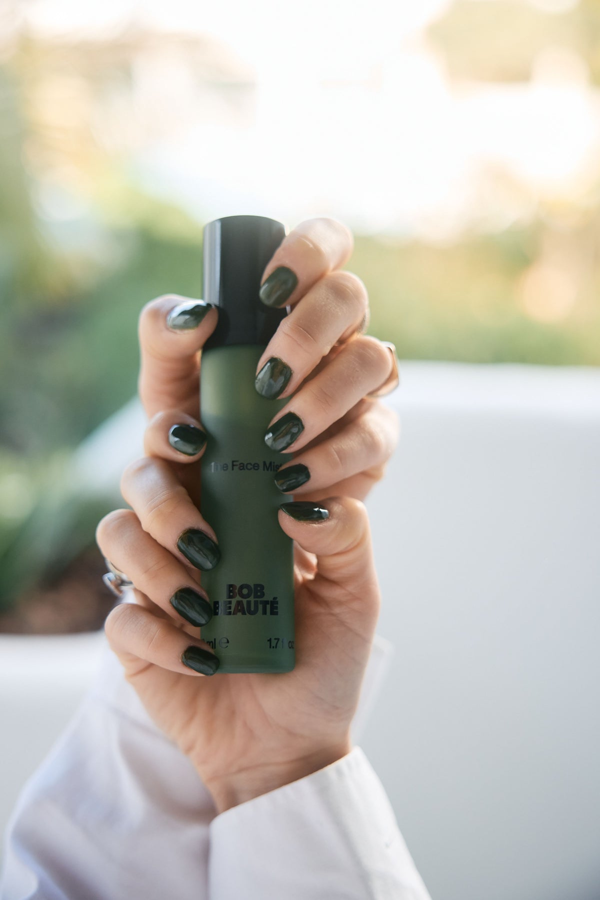 A hand with dark green nails holding a dark green BOB BEAUTÉ bottle with a black pump top, against a blurred outdoor background
